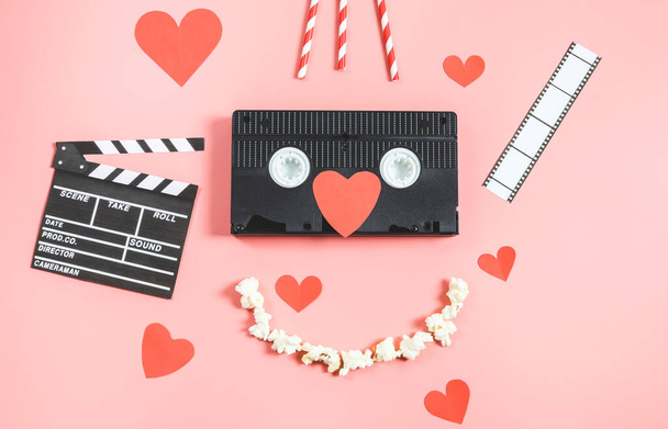 Videószalag, clapperboard, papír szív, film szalag, szívószálak és egy popcorn mosoly fekszik a rózsaszín háttér, felső nézet közelkép. Mozi szerelem és Valentin-napi koncepció. - Fotó, kép