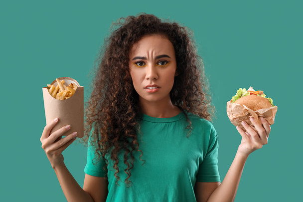 Trieste jonge Afro-Amerikaanse vrouw met frietjes en hamburger op kleur achtergrond - Foto, afbeelding