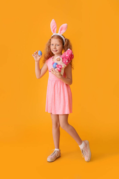 Funny little girl with bunny ears, egg and toy on color background. Easter celebration - Photo, Image
