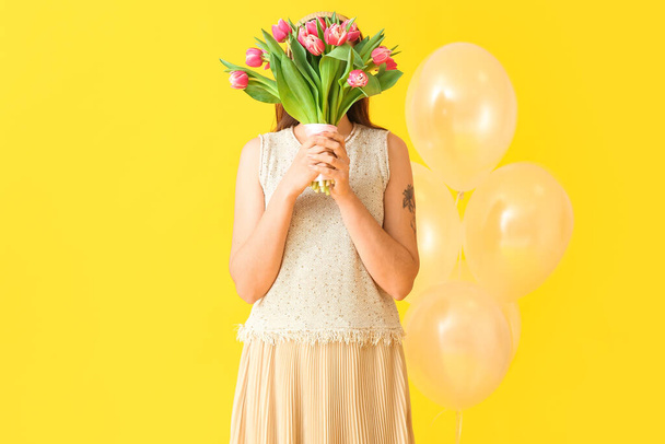 Νεαρή γυναίκα με μπουκέτο τουλίπες και μπαλόνια σε κίτρινο φόντο. Διεθνής Ημέρα της Γυναίκας - Φωτογραφία, εικόνα