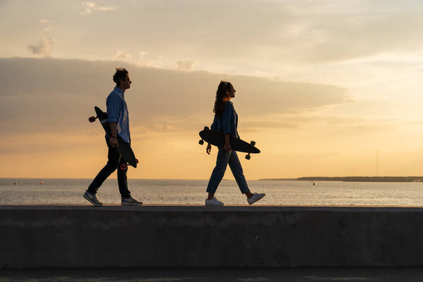 Un paio di giovani longboarder camminano sul mare portando pattini al tramonto. Stile di vita e concetto di libertà - Foto, immagini