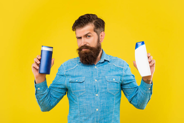 brutal bearded man choose hair conditioner bottle on yellow background, haircare - Foto, Bild