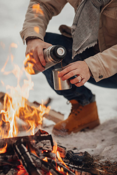 Mężczyzna w zimowym lesie przy ogniu nalewa kawę. Wysokiej jakości zdjęcie - Zdjęcie, obraz