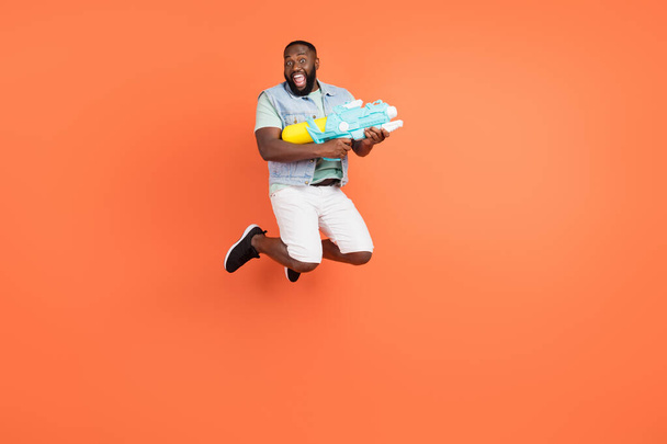 Photo of excited afro guy wear denim vest shooting water gun jumping high isolated orange color background - Foto, Imagem