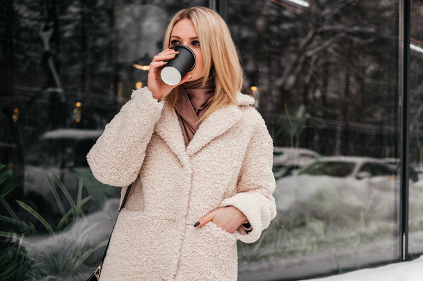Smiling mid age woman drinking coffee outdoors looking away. Caucasian blonde girl in white coat on street. - 写真・画像