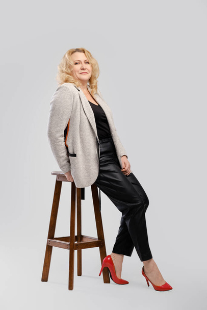 Portrait of senior woman in leather trousers and blazer sitting in profile on over grey studio background - Foto, imagen