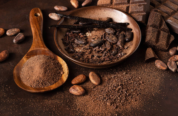 Chocolate and cacao concept. Cocoa powder in bowl near cocoa beans and broken chocolate on black background - Foto, Imagen