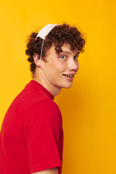 guy with red curly hair wearing a red t-shirt medical mask on the face posing yellow background unaltered - Zdjęcie, obraz
