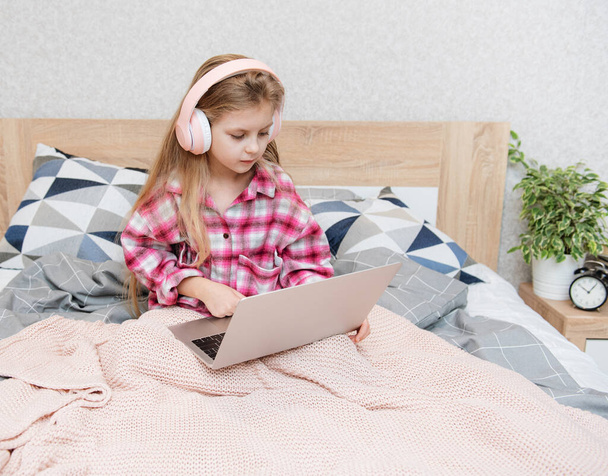 Sonriente niña feliz utilizando el ordenador portátil en los auriculares inalámbricos en la cama en casa. - Foto, imagen