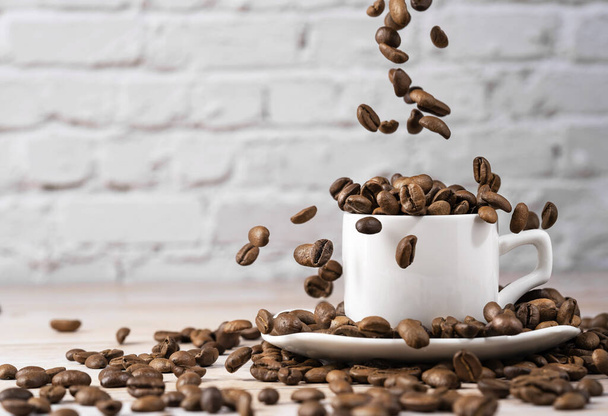 Front view of delicious coffee beans in a cup on wooden table and old gray brick wall background, dark coffee beans flying down - Фото, зображення