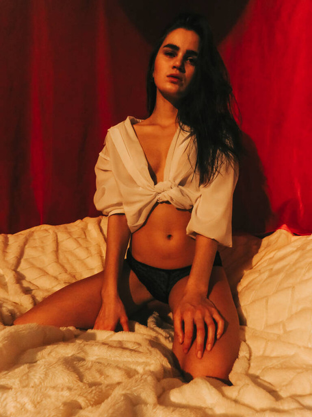 A young brunette woman with dark skin with a slim athletic figure in underwear and a white open shirt sits on a white bedspread on a bed against a background of red curtains, boudoir shooting, the concept of health and beauty of the naked female body - 写真・画像