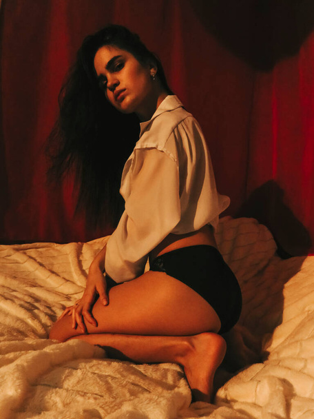A young brunette woman with dark skin with a slim athletic figure in underwear and a white open shirt sits on a white bedspread on a bed against a background of red curtains, boudoir shooting, the concept of health and beauty of the naked female body - Foto, afbeelding