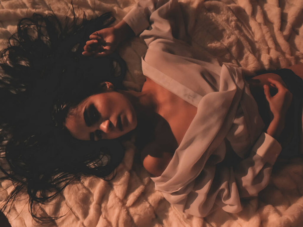 Young sexy brunette woman with a slim athletic figure with loose hair in a white unbuttoned shirt and black panties lying on a light bedspread, top view, boudoir photo shoot, art photo - Φωτογραφία, εικόνα