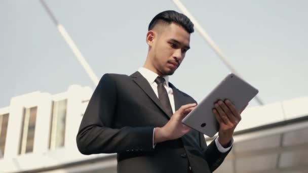 Aantrekkelijke Aziatische zakenman in een zwart pak met behulp van zijn tablet digitaal browsen financiële, scrollen sociale media terwijl staan buiten op straat in de buurt van een groot kantoor gebouw stedelijke. - Video