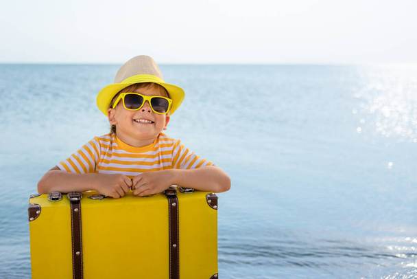 Ευτυχισμένο παιδί που απολαμβάνει τον ήλιο με φόντο τον γαλάζιο ουρανό. Παιδί με βαλίτσα που διασκεδάζει στην παραλία. Καλοκαιρινές διακοπές και διακοπές έννοια - Φωτογραφία, εικόνα