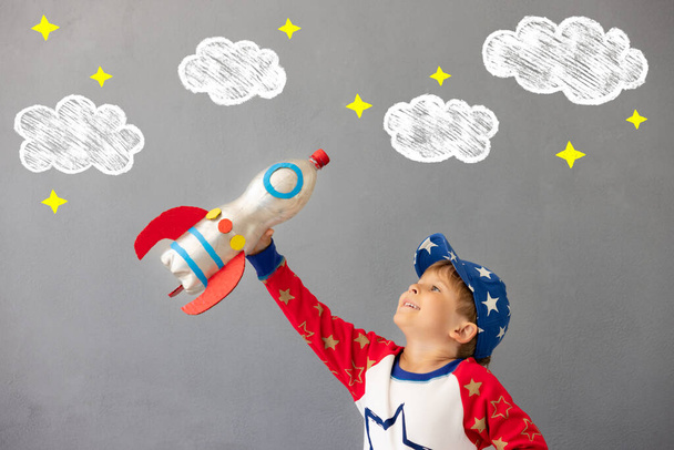 Happy child playing with toy rocket against concrete wall background. Kid pretend to be astronaut. Imagination and children dream concept - 写真・画像