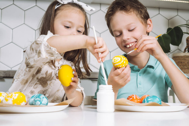 Portrét s úsměvem radostné dvě děti holčička, chlapec, malba, zbarvení velikonoční vajíčka, kuchyňský stůl se štětci. - Fotografie, Obrázek