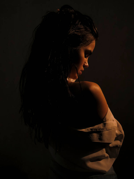 A young half naked brunette woman with dark wet skin in an open white shirt against a gray wall in soft Golden light, the concept of beauty of the female body - Фото, изображение