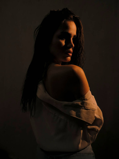A young half naked brunette woman with dark wet skin in an open white shirt against a gray wall in soft Golden light, the concept of beauty of the female body - 写真・画像