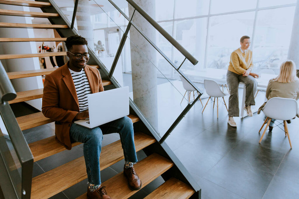 Schöner junger afroamerikanischer Geschäftsmann arbeitet am Laptop, während er auf einer Bürotreppe sitzt - Foto, Bild