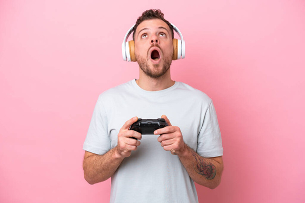 Young Brazilian man playing with video game controller isolated on pink background looking up and with surprised expression - Photo, Image