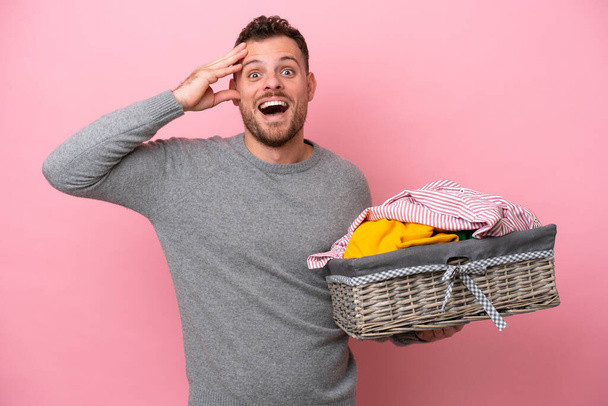 Junger brasilianischer Mann mit einem Wäschekorb auf rosa Hintergrund und überraschendem Ausdruck - Foto, Bild