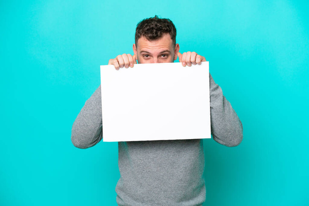 Young Brazilian man holding an empty placard isolated on blue background holding an empty placard and hiding behind it - Photo, image