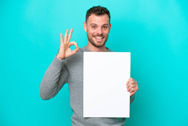 Junger brasilianischer Mann hält ein leeres Plakat auf blauem Hintergrund, hält ein leeres Plakat und tut OK-Zeichen - Foto, Bild