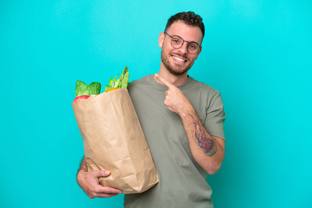 Jonge Braziliaanse man met een boodschappentas geïsoleerd op blauwe achtergrond wijzend naar de zijkant om een product te presenteren - Foto, afbeelding