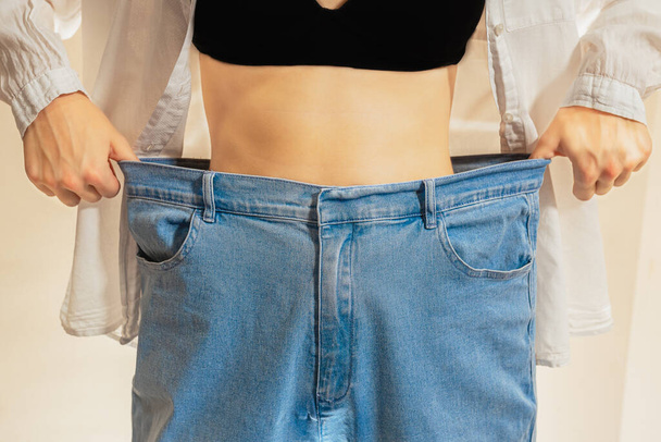 Štíhlá žena v širokých džínách. Oříznutý záběr mladé štíhlé ženy stojící s nahým břichem v širokých džínách a černou střechou ukazuje úspěšný hubnutí dieta a wellness koncept - Fotografie, Obrázek