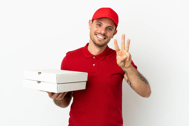 dostawca pizzy z mundurkiem roboczym podnosząc pudełka po pizzy na odizolowanej białej ścianie szczęśliwy i licząc trzy palcami - Zdjęcie, obraz