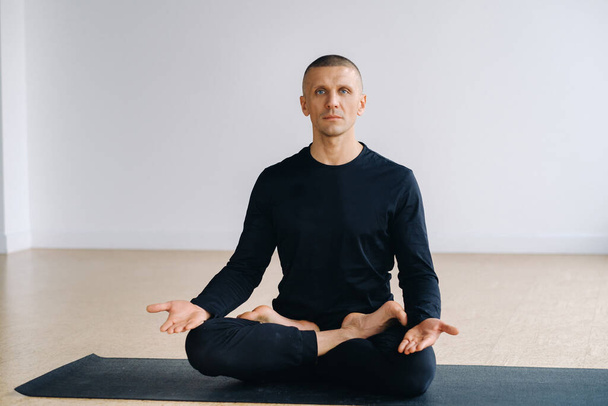 A male athlete in black sportswear is sitting on a mat in the lotus position in the gym. - Foto, Imagem