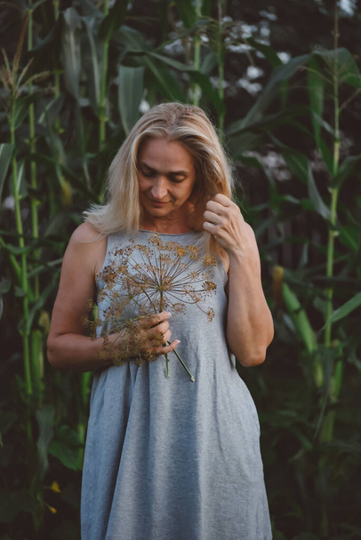 Photo in motion, a woman with short blond hair in a gray summer dress posing outdoors with a branch of dill in her hands. The girl poses against the backdrop of a corn field. The concept of the garden and vegetable garden. - Fotografie, Obrázek