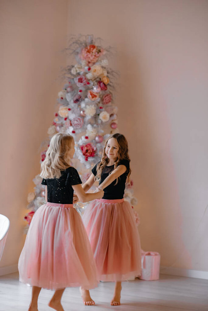 Two smiling little girls are posing by the Christmas tree. A child in a New Year's location. Photo studio with New Year's decorations. New Year's gifts, white tree. Merry Christmas. Selective focus. - Фото, изображение