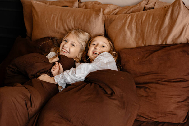 In motion, a portrait of two little happy girls lying on the bed in the morning, the children woke up in bed in the morning in a good mood. Good morning. Children in pajamas. Carefree childhood. - Foto, imagen