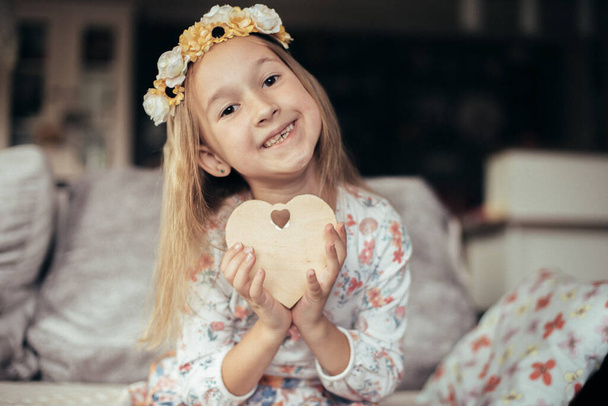Portrait of a cute smiling little girl in a white shirt with a wreath of flowers on her head with a heart in her hands. Wooden craft made of wood in the form of a heart in a child's hands. - 写真・画像