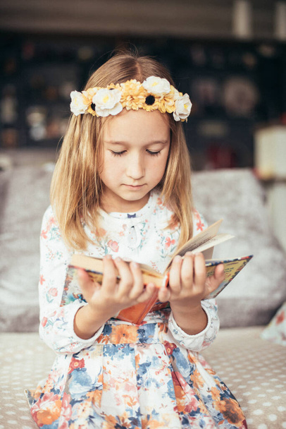 Dítě si čte knihu, zatímco sedí v místnosti. Portrét roztomilé holčičky s věnečkem květin na hlavě a knihou v rukou. Děvče si dělá úkoly. Koncept učení a znalostí. - Fotografie, Obrázek