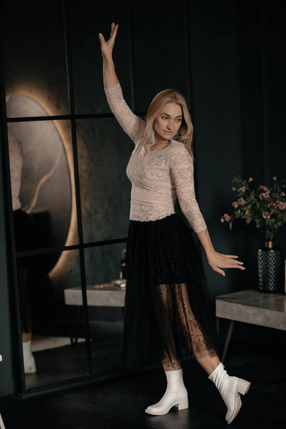 Photo in motion, a girl with short blond hair in a black skirt and white high-heeled shoes dances in a dark studio near a mirror. A beautiful girl is dancing. The concept of fun and dancing, disco. - Фото, изображение