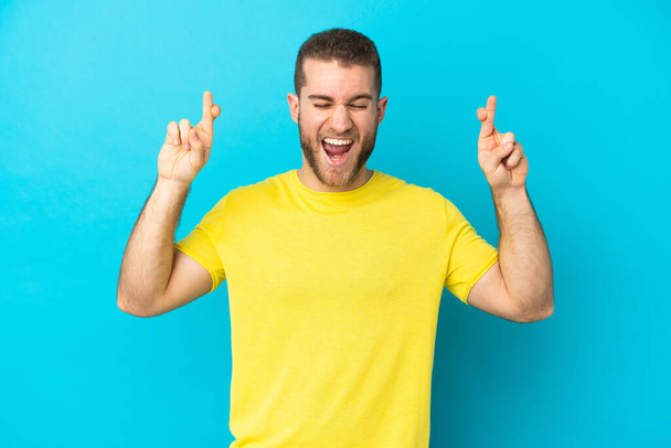 Young handsome caucasian man isolated on blue background with fingers crossing - Foto, imagen