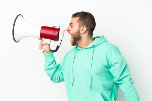 Homem caucasiano bonito jovem isolado em fundo branco gritando através de um megafone para anunciar algo em posição lateral - Foto, Imagem