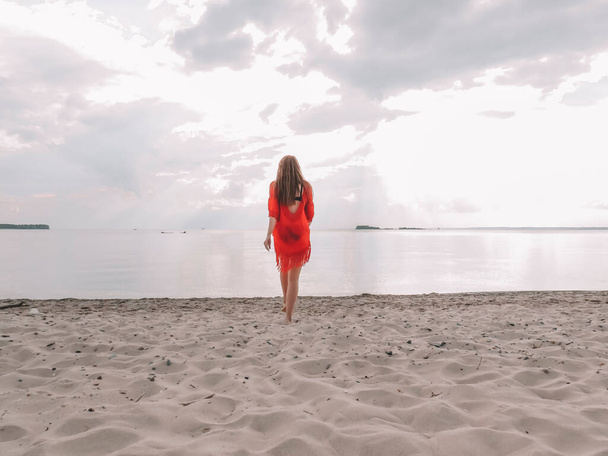 Young  woman with loose red hair in red beach dress on the sand of the beach, concept of outdoor recreation, beach season, female beauty, harmony and unity with nature - 写真・画像