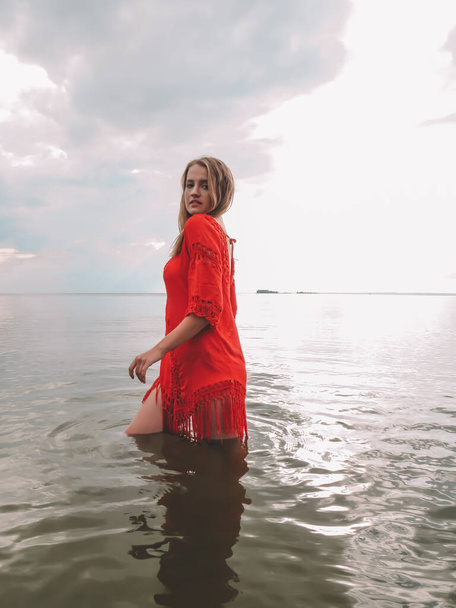 Young woman with loose red hair in a red beach dress in the water in the shallow waters of the sea bay on a hot summer day, the concept of summer vacation at sea, beach season, vacation/ Sunday weekend in nature, sun tanning - Photo, image