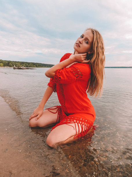 Young blonde woman in short red dress sitting in the water on the shallows on the sandy beach of the sea on a summer sunny day, the concept of a healthy lifestyle, outdoor activities, beach season, weekend and vacation at the sea - Φωτογραφία, εικόνα