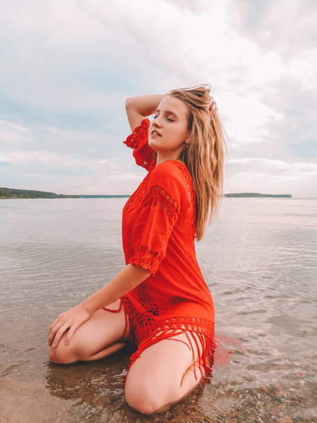 Young blonde woman in short red dress sitting in the water on the shallows on the sandy beach of the sea on a summer sunny day, the concept of a healthy lifestyle, outdoor activities, beach season, weekend and vacation at the sea - Foto, Imagem