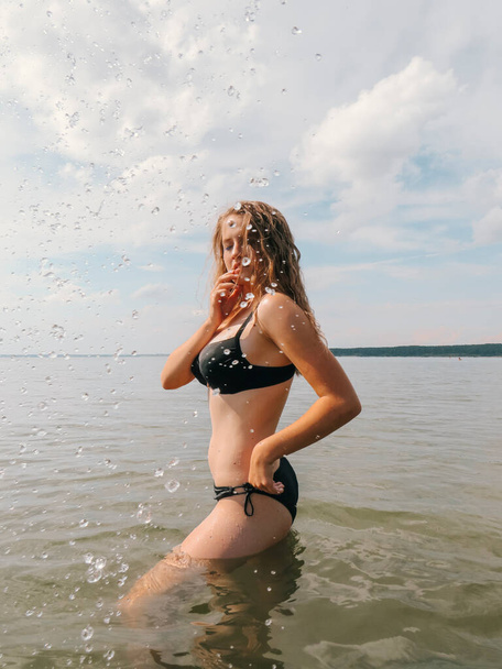 Beautiful young woman with a slender figure on the background of water splashes on the shallow waters of the sea beach on a hot summer day, the concept of beach holidays, sun tanning, swimming - 写真・画像