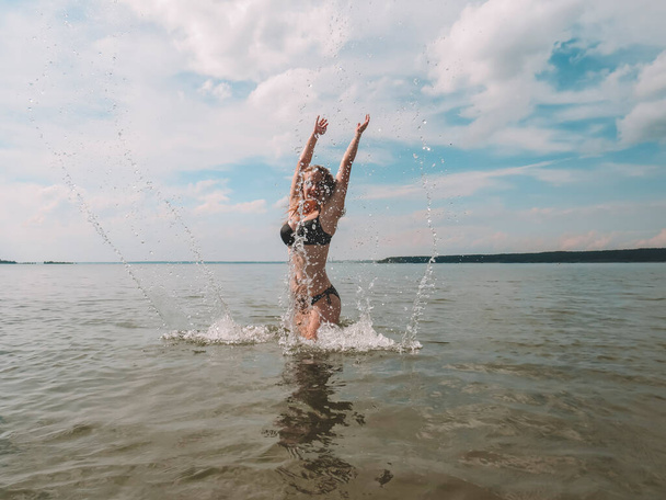 Beautiful young woman with a slender figure on the background of water splashes on the shallow waters of the sea beach on a hot summer day, the concept of beach holidays, sun tanning, swimming - Fotografie, Obrázek