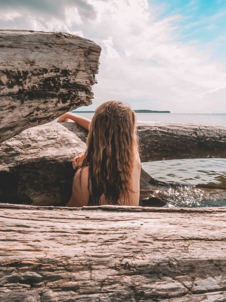 Red-haired young woman with loose hair with a naked beautiful body in the water of the sea bay at the trunk of a huge dry tree on a summer sunny day, art photo, image of a mermaid, close-up. The concept of beauty and health,beach holidays,sun tanning - Foto, Imagem