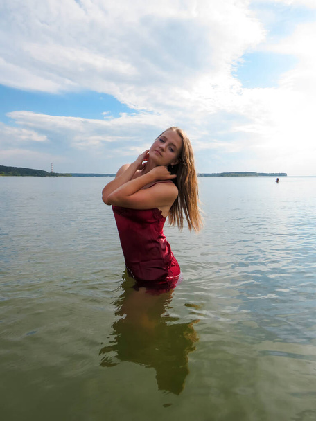 Attractive young blonde woman with loose hair and sexy slender body in a tight wet red silk dress in the water on a shallow beach on a hot summer day, sun glare on the water, the concept of a beach holiday, sun tanning and swimming in the sea - Foto, Imagem