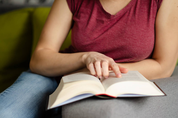 Selective focus on book read by caucasian woman in burgundy T-shirt and jeans sitting on gray sofa with pillows. Concept of rest in cozy house. Time alone with yourself. - 写真・画像