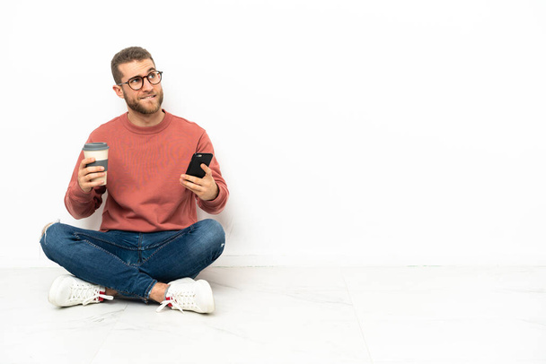 Jeune bel homme assis sur le sol tenant du café à emporter et un mobile tout en pensant à quelque chose - Photo, image